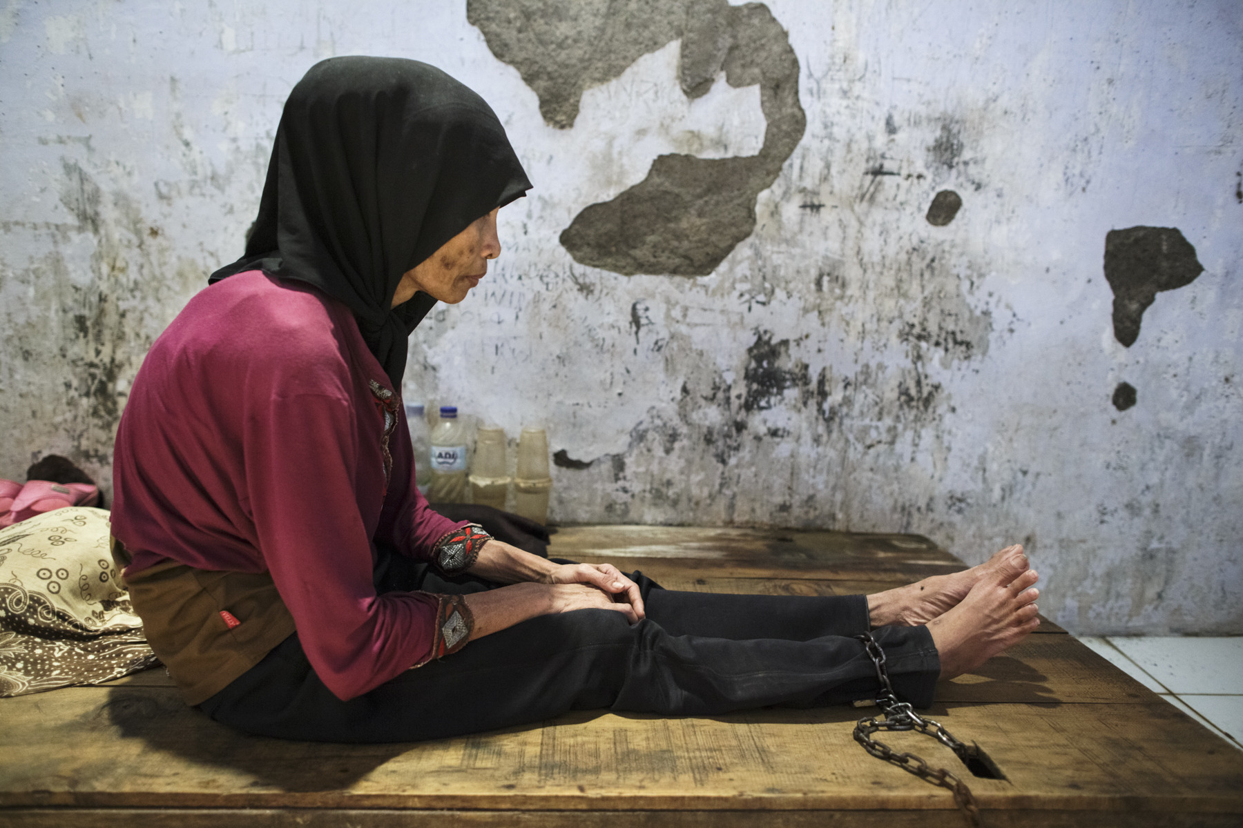 A young woman with a real or perceived psychosocial disability sits with her ankle chained to a platform bed at Bina Lestari faith healing center in Brebes, Central Java. Photo: &copy; 2018 Andrea Star Reese